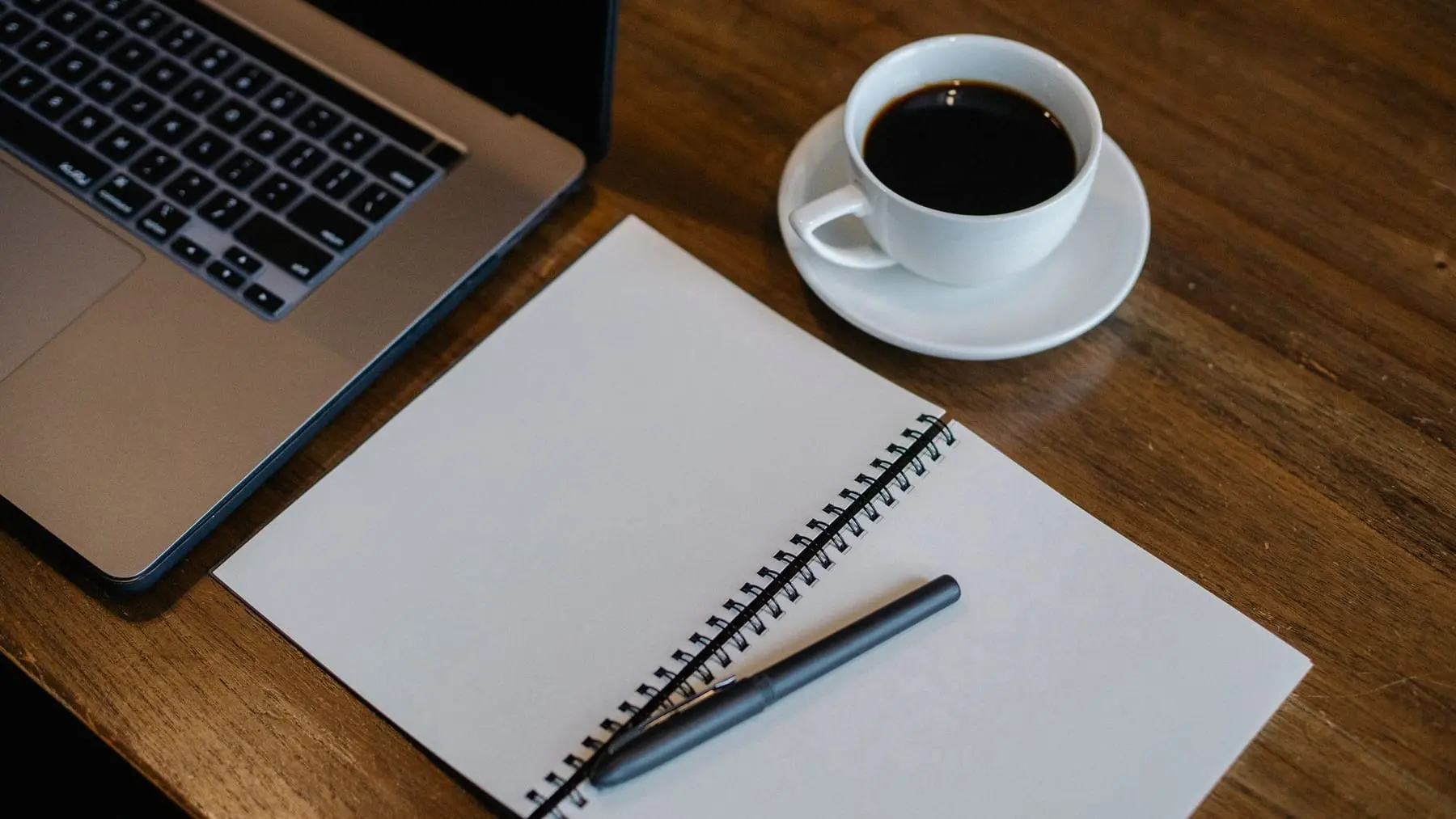 A notebook, a laptop and a cup of coffee on a wooden desk